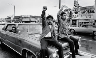 Two young Chicano men ride on the hood of a car and raise their fists during a National Chicano Moratorium Committee march in Los Angeles, California, on August 29, 1970, in opposition to the war in Vietnam.