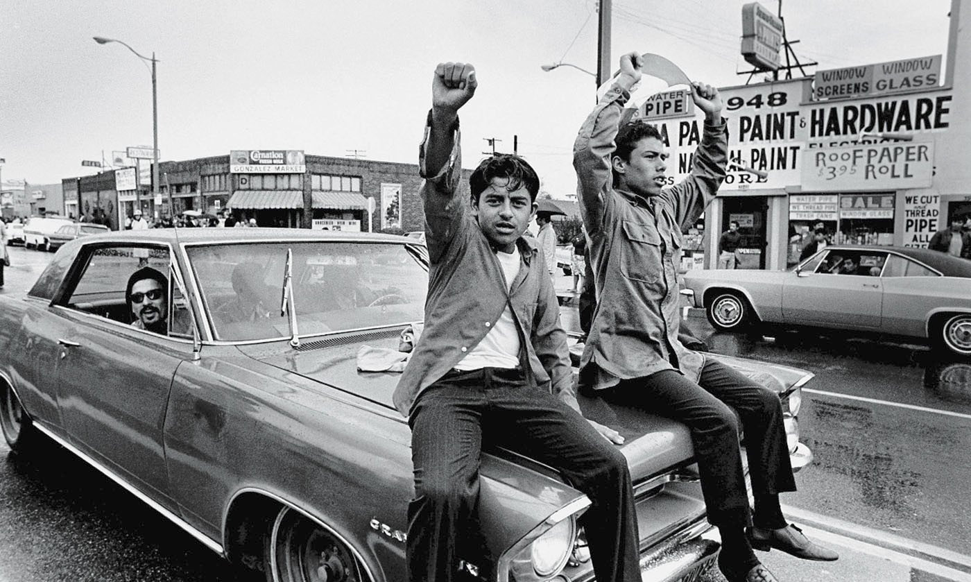 Two young Chicano men ride on the hood of a car and raise their fists during a National Chicano Moratorium Committee march in Los Angeles, California, on August 29, 1970, in opposition to the war in Vietnam.