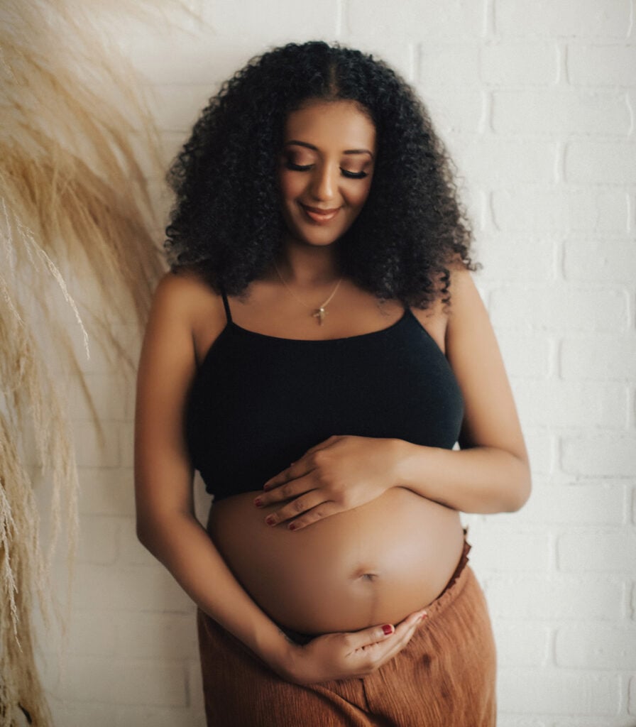 Gloria Alamrew, 30 weeks pregnant, cradles her pregnant belly with her hands. She wears a black tank top, lifted to reveal her belly, and brown flowing pants, while standing against a white brick wall. 