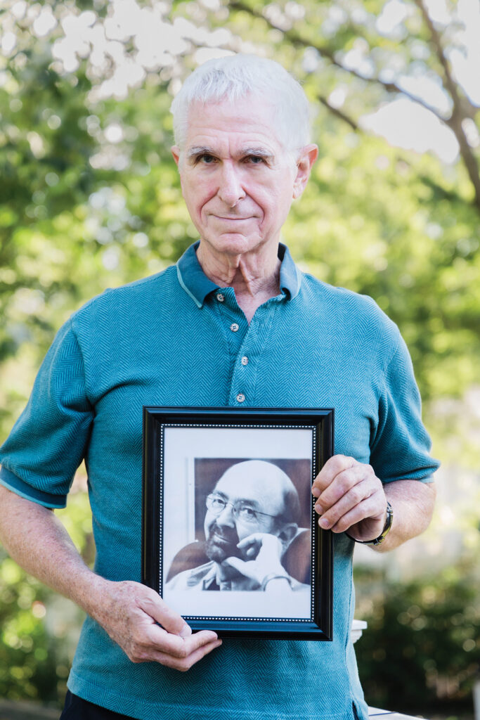 Charles Koehler, wearing a blue polo shirt, holds a black-and-white photo of his late husband, Dennis Hostetler, at age 68. He is standing outdoors, with trees and foliage visible in the background. 