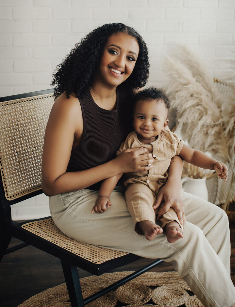 Gloria Alamrew sits on a black and wicker woven chair, with her 10-month-old son sitting on her lap. Both are looking directly into the camera and smiling. 