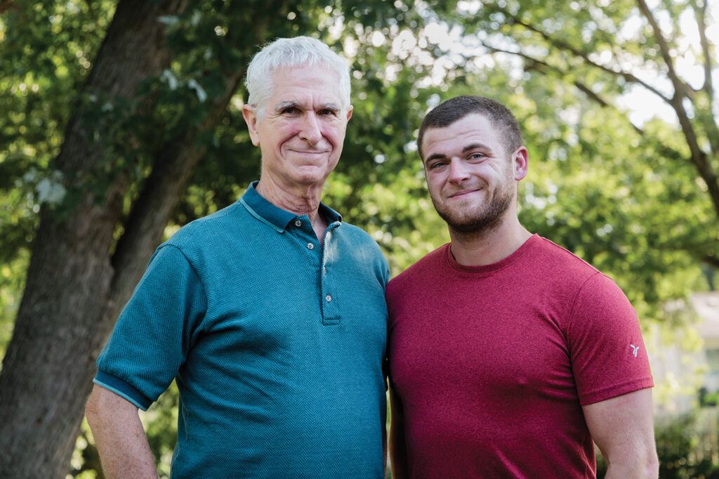 Charles Koehler, in a blue polo shirt, stands next to his son Spencer, age 28, wearing a red T-shirt. Both men are smiling, and standing outdoors with trees and foliage visible in the background. 