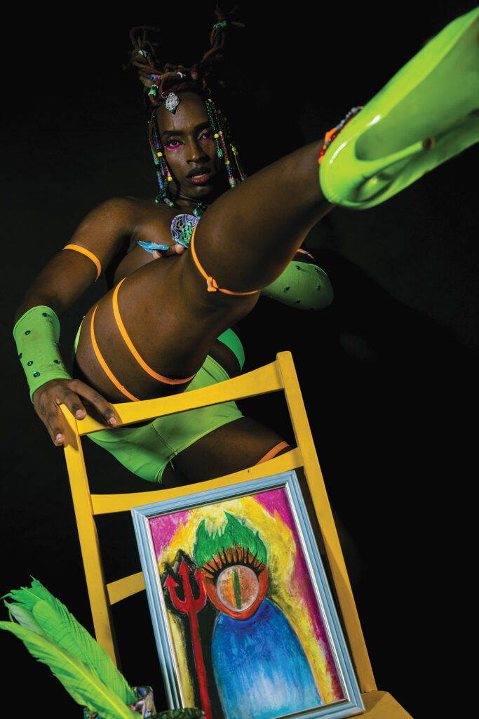 Puma Camillê stands against an all-black background, wearing bright green high heels, with matching bejeweled gauntlets and shorts. Her leg is lifted straight out toward the camera, above a yellow chair on which an abstract painting of an eye and pitchfork sit.