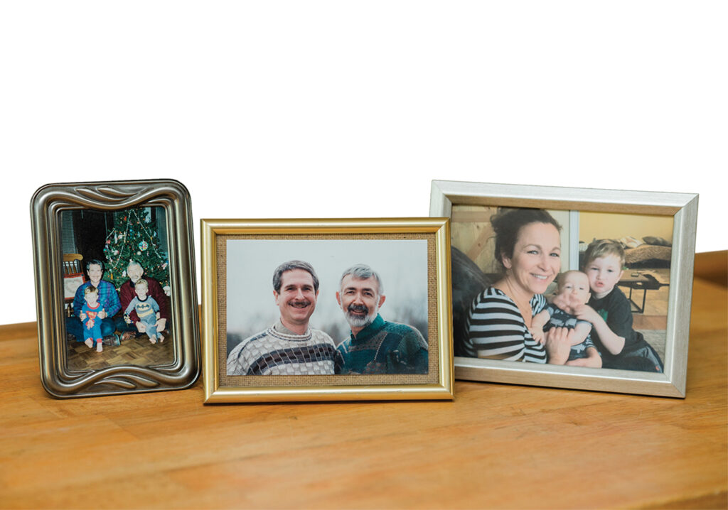 Three framed photographs sit on a wooden shelf. The photos show, from left: Charles, age 45 (left) with Dennis, age 55 (right) holding their sons Connor, age 2 (left) and Spencer, age 4 (right) on Christmas 1997, in front of a decorated Christmas tree. Center: Charles, age 45, with Dennis, age 55; both wearing patterned sweaters and smiling broadly. Right: Jocelyn, Dennis’ daughter from a previous 12-year marriage, smiles while holding her two children; one an infant, and one a toddler with their arms around their sibling.