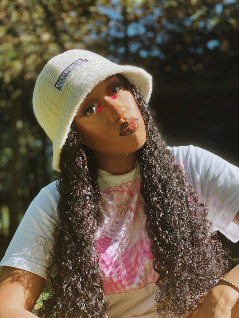 Zalika U. Ibaorimi is seated outdoors, looking directly into the camera. She wears a plush yellow bucket hat, a white t-shirt with a pink pointillism pattern, and her long, curly dark brown hair hangs down to her wrists.