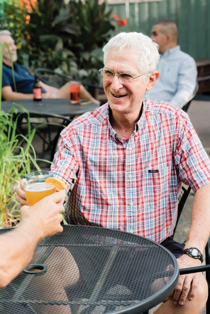 Charles shares drinks with members of his local PrimeTimers social group, of which he was founding president, on the back patio of Just John Night Club in St. Louis, Missouri. He wears a red, white, and blue-checked shirt, glasses, and holds up a pint of beer to toast with a friend who is off-camera.