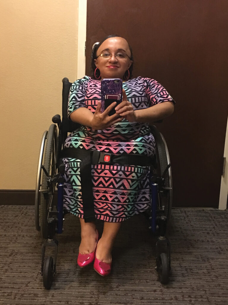 Vilissa Thompson takes a selfie in a room with grey carpet and tan walls. She is seated in her wheelchair, and wearing glasses, pink hoop earrings, and a full-length dress patterned with black geometric shapes on top of vertical bands of neon pink, purple, and turquoise.