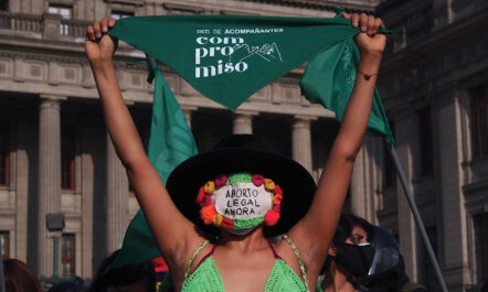 A pro-abortion activist, holding a green bandana aloft, and wearing a black hat and a handmade mask reading “legal abortion now” in Spanish, participates in a September 2021 protest in Lima, Peru.
