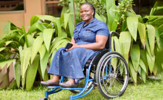 Betty, a Black woman wearing a denim dress sitting on a blue wheelchair, smiles for the camera. There's bright green foliage in the background.