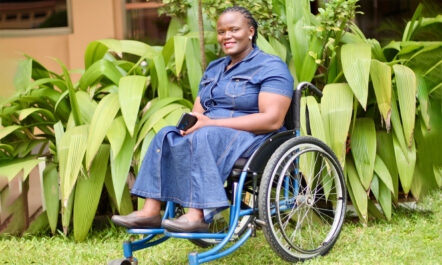 Betty, a Black woman wearing a denim dress sitting on a blue wheelchair, smiles for the camera. There's bright green foliage in the background.