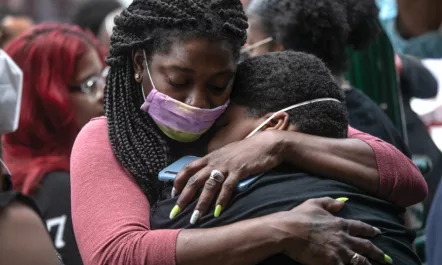 A Black woman, with dreadlocks and wearing a pink mask, hugs another Black person at the funeral for Conrad Coleman Jr., on July 3, 2022 in New Rochelle, New York.