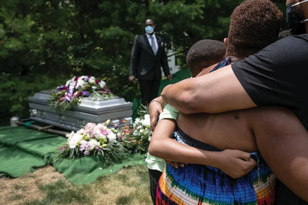 Three Black people embrace while overlooking the silver casket of Conrad Coleman Jr., which is sitting above the burial plot, adorned with flowers. 