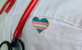 Photo of trans flag colored heart on a doctor's pin.
