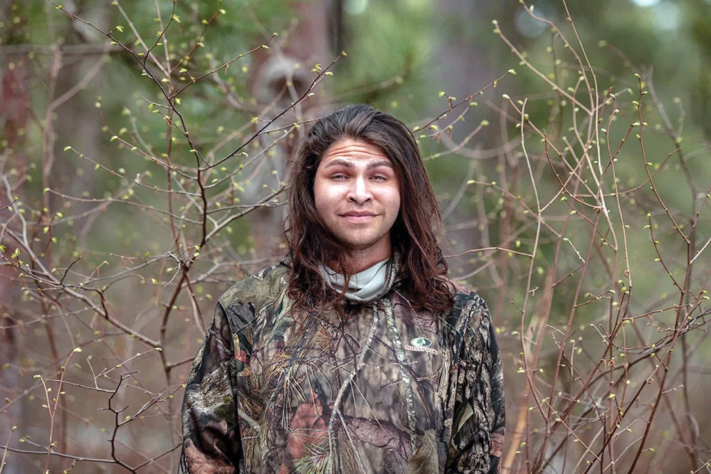 Big Wind Carpenter stands outdoors, surrounded by sparse branches with small green buds. They are looking directly at the camera, and wearing a hoodie with a forest camouflage pattern.