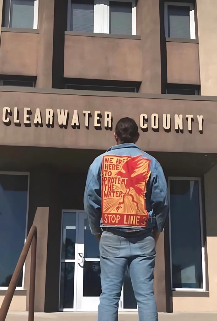 Big Wind Carpenter is pictured standing in front of the Clearwater County Courthouse in Bagley, Minnesota. Their back is to the camera, and they are wearing all denim, including a jacket with a large yellow patch on the back with red lettering reading "We are here to protect the water. Stop Line 3."