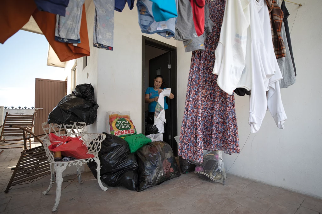 Elva Rivas stands in the doorway of her home, sorting secondhand clothing that she sells at local markets every weekend to make ends meet.