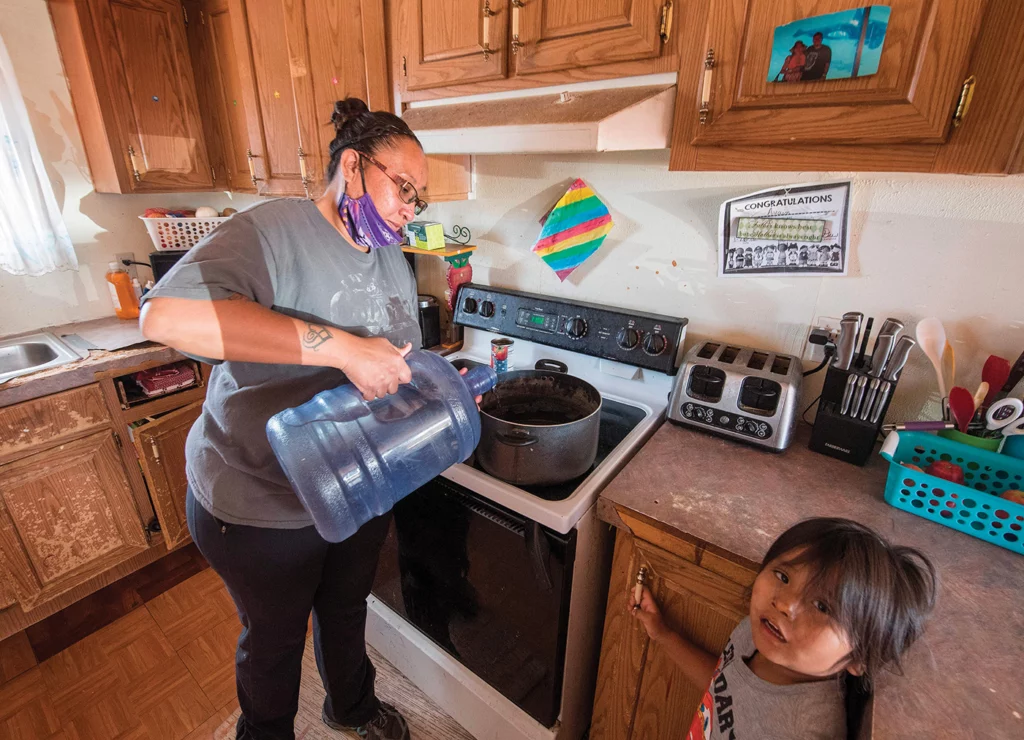Amanda Larson pours bottled water into a pot on her stove to bathe her son, Gary Jr., in their home in Thoreau, New Mexico, on May 22, 2020. Gary Jr. is looking up at the camera, while Larson, with a purple mask pulled down around her chin, focuses on the water.