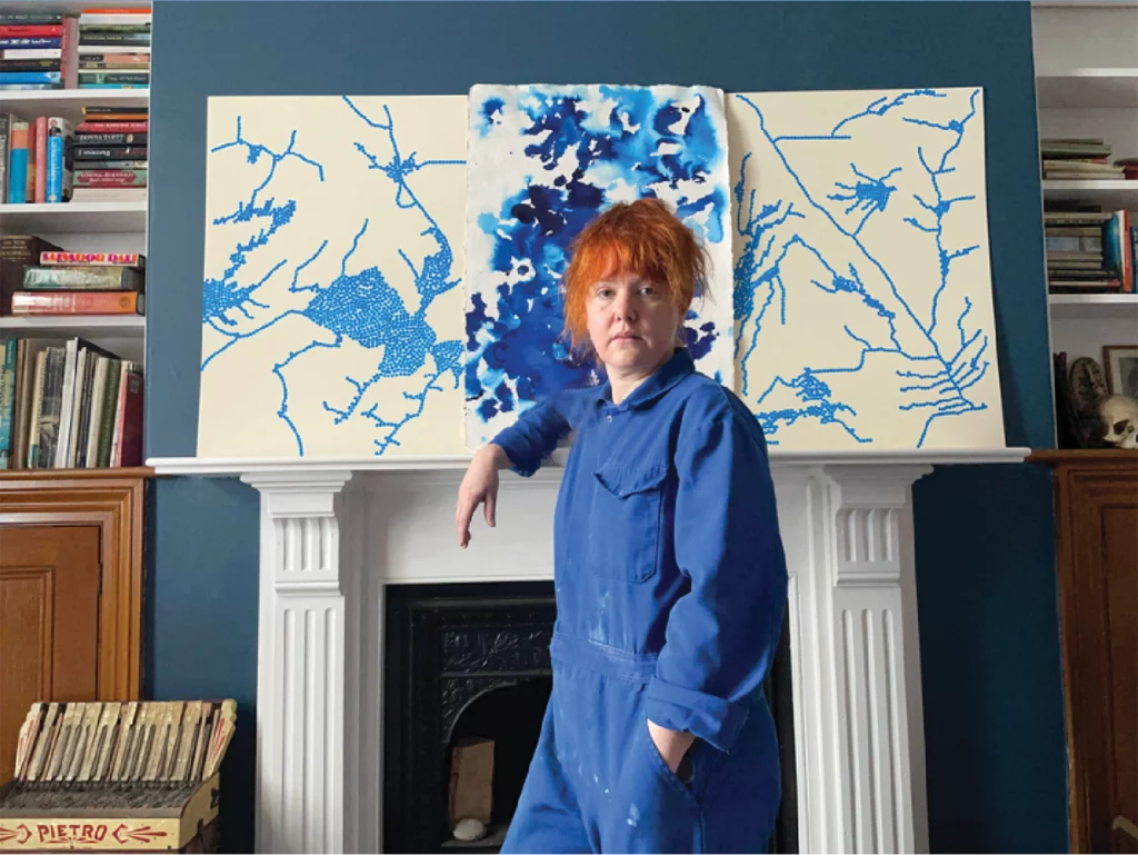 Rebecca Wyn Kelly stands in front of a fireplace, which features her artwork on the mantle. Kelly's jumpsuit matches the shade of blue used in her paintings.