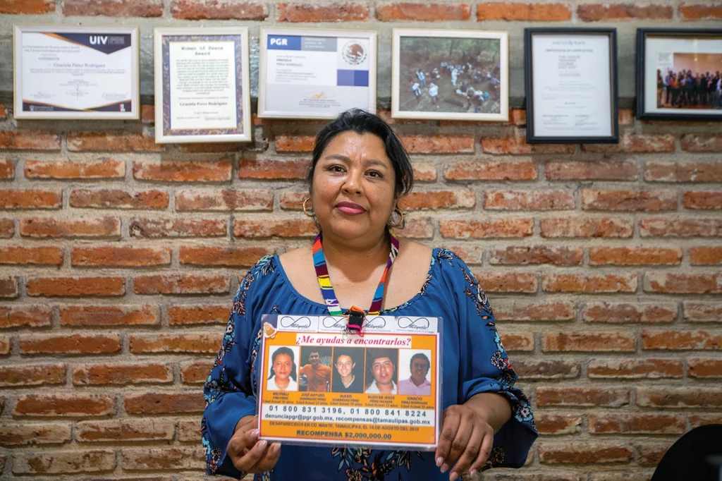 Graciela Pérez stands in front of a brick wall, holding a missing person sign showing her disappeared loved ones, at the office of Milynali Red AC, an organization she co-founded—and named after her disappeared daughter—to advance efforts to find and locate disappeared people in Mexico.