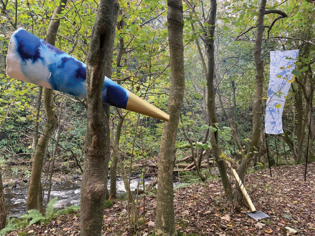 This photo of Rebecca Wyn Kelly's "listening cones" depicts large blue and gold paper cones set amongst trees near a river. A white and blue patterned banner is visible hanging from trees in the background.