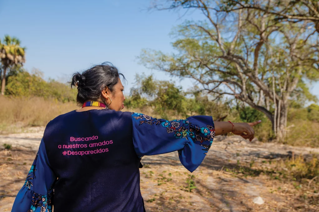 Graciela Pérez points to the location of an “extermination site” identified by government workers at a property in Tamuín, San Luis Potosí, Mexico. She is wearing a blue vest with pink text on its back, reading "Buscando a nuestros amados #desaparecidos" ("Searching for our loved ones #disappeared")