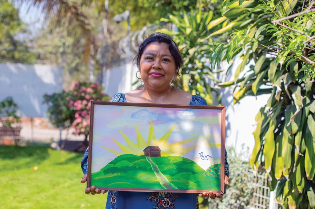 Graciela Perez stands outside in a garden, holding a painting by her daughter before she disappeared at age 13. The painting depicts a brown house atop a green hill, surrounded by sunbeams and colorful clouds.