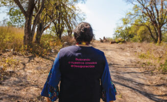 Graciela Pérez is photographed from the back, walking down a sandy road lined with light green trees and sparse grasses in Tamuín, San Luis Potosí, Mexico. She wears a vest that reads, in Spanish, "Searching for our loved ones #disappeared"
