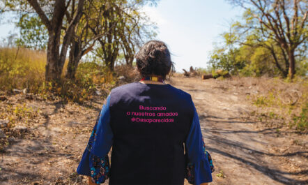 Graciela Pérez is photographed from the back, walking down a sandy road lined with light green trees and sparse grasses in Tamuín, San Luis Potosí, Mexico. She wears a vest that reads, in Spanish, "Searching for our loved ones #disappeared"