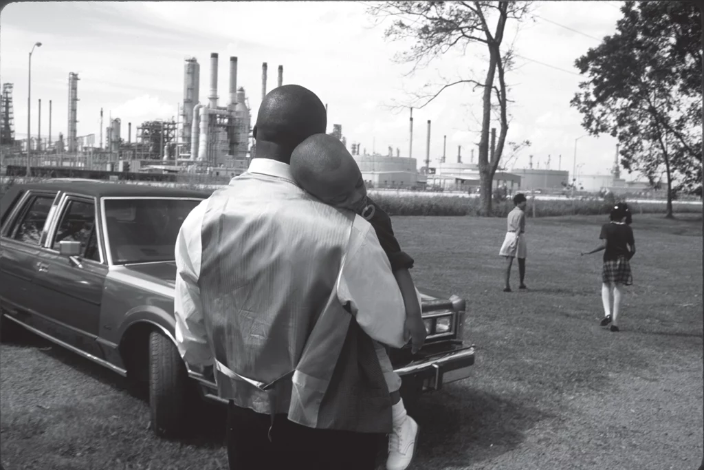 A family leaves Sunday church services in Lions, Louisiana, in October 1998. In the foreground, a man holds a sleeping child on his shoulder, standing in front of a car, with two women visible in the midground. An industrial chemical plant looms in the background.