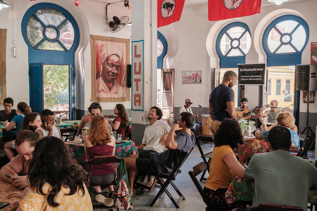 A light, airy restaurant space with white walls and ornate windows and doors with blue trim, is filled with tables occupied by diners enjoying a three-course menu made from food produced by Roseli Nunes families. A three-piece band can also be seen playing at the front of the room.