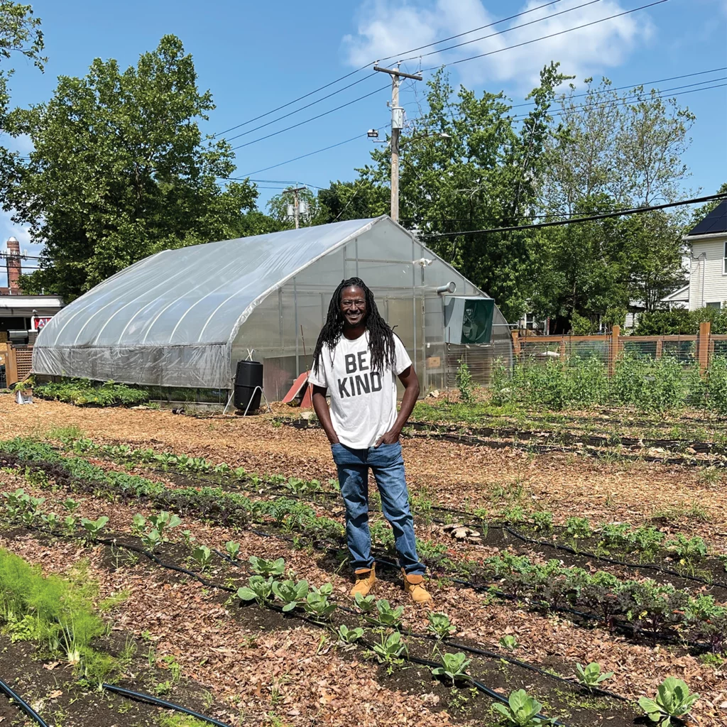 Jonathón Savage stands among growing seedlings at the Gather New Haven farm. He wears a shirt reading "Be Kind" and there is a greenhouse in the background.