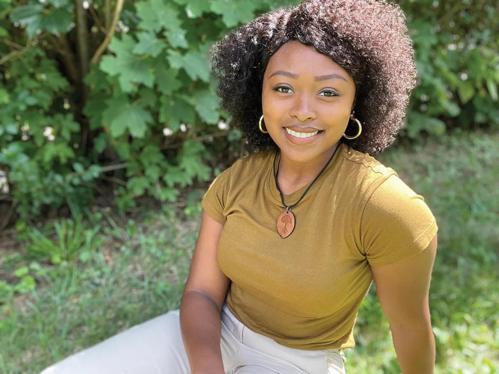 Vetivieah Harrison smiles at the camera, seated on grass with green foliage in the background.