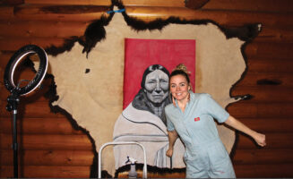 Artist Miah Chalfant, a light-skinned Native artist, smiles and poses in front of one of her tallow-hide paintings: a black-and-white portrait of a tribe matriarch in front of a deep, red square, all on a large, tan hide.