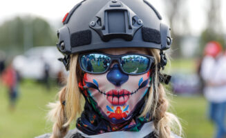 A close-up photo of a white, blonde female protester in Portland, Oregon. She is wearing a protective helmet, sunglasses, and a face mask concealing her mouth and nose.