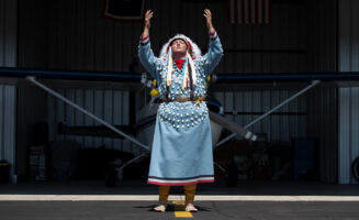 Shawnee Real Bird, in full regalia, poses in front of her airplane in a hangar. She is raising both her hands and looking skyward.