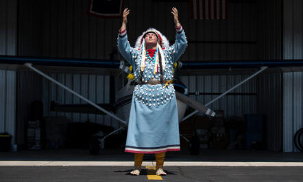 Shawnee Real Bird, in full regalia, poses in front of her airplane in a hangar. She is raising both her hands and looking skyward.