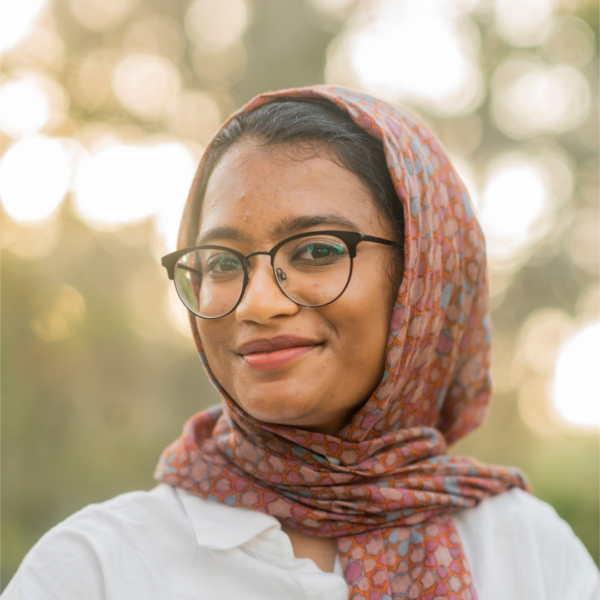 Amal Ahmed, a Muslim woman wearing a hijab, looks at the camera. She is wearing glasses.