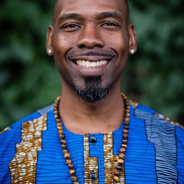 a Black man wearing a blue shirt and necklaces smiles at the camera