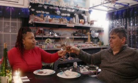 An image from the documentary Patrice: The Movie, which is available to stream on Hulu. Patrice, a black woman wearing a red shirt, has a toast with wine glasses with her partner Gerry, a white man in a grey sweater, both seated at a dinner.