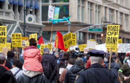 A view from behind of people marching in New York City carrying signs with messages like 'Hands off Iran' and 'No new US war in the middle east.' A police officer watches in the foreground.