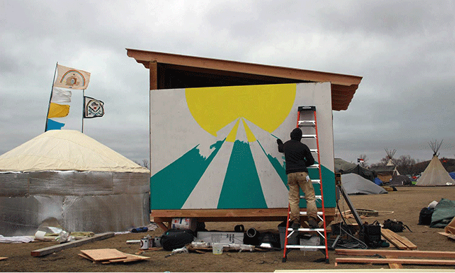 Roger Peet paints the side of one of the cabins reassembled at the Standing Rock encampment. Photo by Roger Peet.